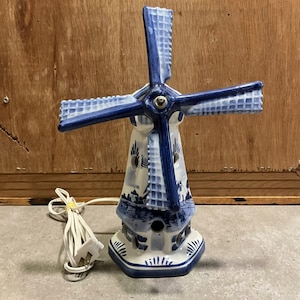 Może przedstawiać: Ceramiczna lampa w kształcie wiatraka w kolorze niebiesko-białym, z szczegółowym wzorem holenderskiego krajobrazu. Lampa posiada czteropłatowy wentylator i podstawę z dekoracyjnymi wycięciami. Przewód elektryczny i wtyczka są białe.