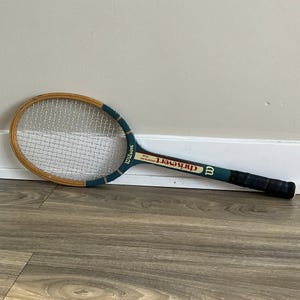 Puede incluir: Raqueta de tenis Wilson Chris Evert American Star vintage. La raqueta tiene un marco de madera con un mango y cabeza verde azulado y marrón. El agarre es negro y las cuerdas son blancas. Presenta el logotipo de Wilson y el nombre de Chris Evert.