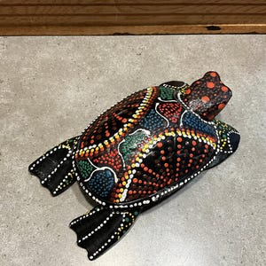 Puede incluir: Figura de tortuga de madera pintada a mano. El caparazón de la tortuga es negro con un colorido patrón de puntos en rojo, azul, verde y amarillo. La cabeza y las aletas son negras con detalles de puntos blancos. Un artículo decorativo.