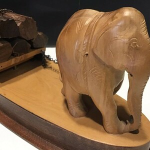 May include: A wooden elephant figurine pulling a small wooden cart loaded with logs. The elephant is carved with intricate details and has a realistic appearance. The cart is on wheels and has a chain connecting it to the elephant.