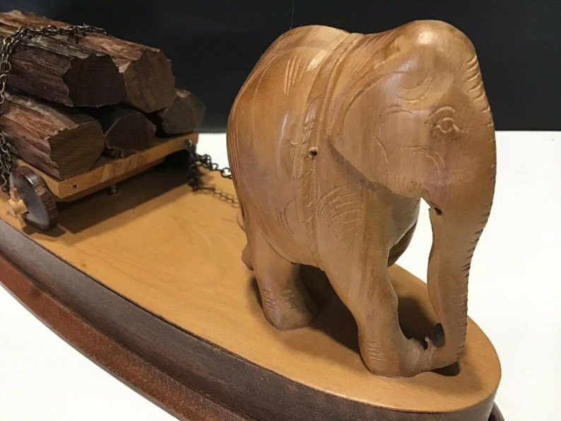 May include: A wooden elephant figurine pulling a small wooden cart loaded with logs. The elephant is carved with intricate details and has a realistic appearance. The cart is on wheels and has a chain connecting it to the elephant.