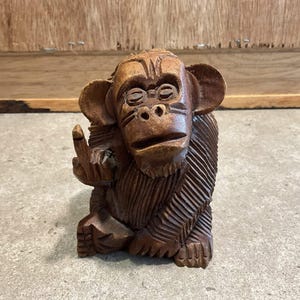 May include: A carved wooden monkey figurine in shades of brown. The monkey is seated and giving the middle finger. The sculpture has detailed carving, showing fur and facial features. The background is a wooden surface.