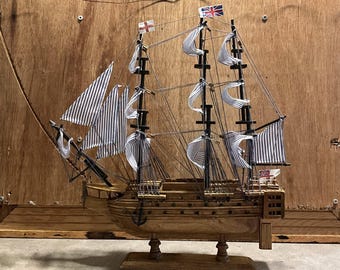 May include: A wooden model of a tall ship with white and blue striped sails and multiple masts. The ship has several flags, including the Union Jack and the English flag. The model is mounted on a wooden base.