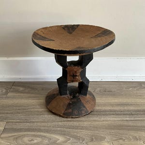 Puede incluir: Un taburete de madera con una parte superior redonda y plana y una base tallada. La parte superior está pintada de marrón con diseños geométricos negros. La base está pintada de negro con un diseño geométrico marrón.
