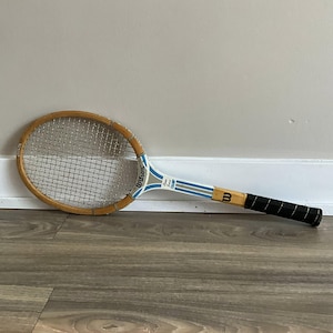 Puede incluir: Raqueta de tenis Wilson vintage con marco de madera y malla plateada. El mango es negro con un diseño blanco y azul en el eje. La raqueta está sobre una superficie de madera.