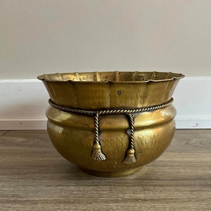 May include: A large, gold-toned brass planter with a scalloped rim and a rope-like design around the top. The planter has two tassels hanging from the rope.