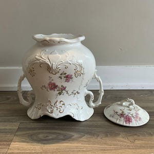 May include: A white ceramic urn with a lid, featuring floral designs in pink and gold accents. The urn has two handles and ornate detailing. The lid mirrors the floral pattern. A decorative piece for home decor.