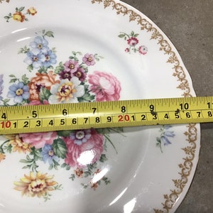 May include: White ceramic dinner plate with a gold trim and a floral design featuring pink, yellow, blue, and purple flowers. The plate is 11 inches in diameter.