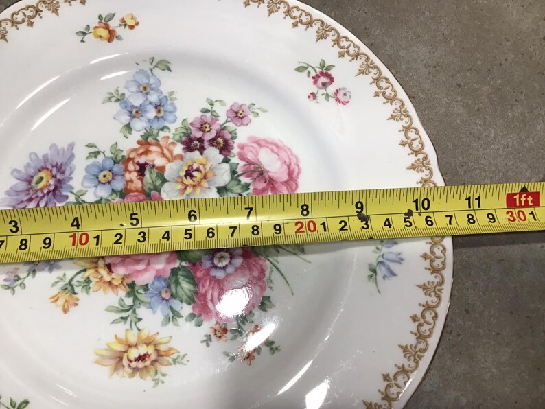 May include: White ceramic dinner plate with a gold trim and a floral design featuring pink, yellow, blue, and purple flowers. The plate is 11 inches in diameter.