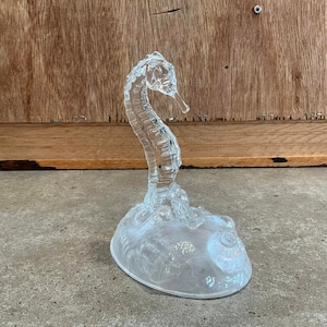 May include: A clear glass seahorse figurine with a detailed design. The seahorse is standing on a base with a seashell design.
