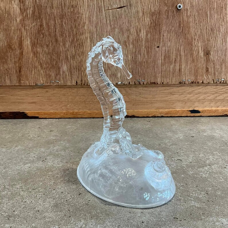 May include: A clear glass seahorse figurine with a detailed design. The seahorse is standing on a base with a seashell design.