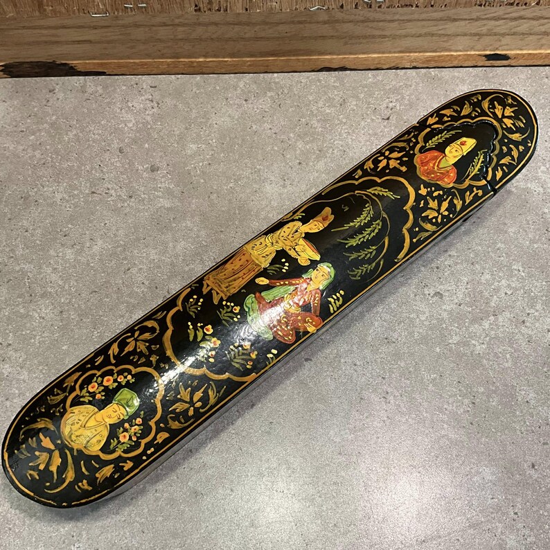 May include: A long, black, decorative box with a lid. The box is adorned with colorful illustrations of figures and floral patterns in gold, red, and green. The box is likely used for storage or display.