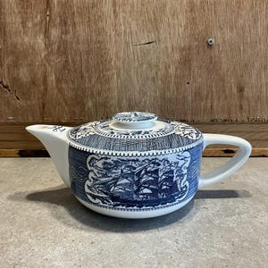 May include: A white teapot with a blue and white nautical design. The teapot has a round body and a short spout. The design features a ship with sails and a rope.