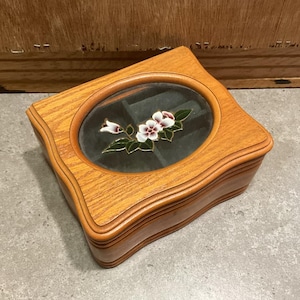 May include: A wooden jewellery box with a glass top featuring a floral design. The box has a wavy edge and a compartment for storing jewellery.