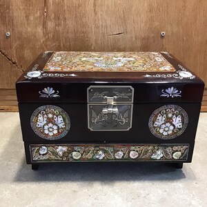 Può includere: Una scatola per gioielli in legno marrone scuro con intricati disegni di intarsi in madreperla. La scatola ha un coperchio incernierato e una chiusura in metallo decorativa. L'intarsio presenta motivi floreali, farfalle e disegni geometrici.