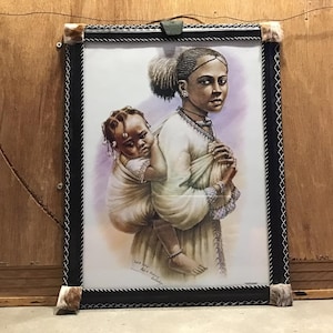 May include: Framed art print of a woman carrying a child on her back. The woman is wearing a white dress and the child is wearing a white garment. The artwork is in a black frame with a silver border.