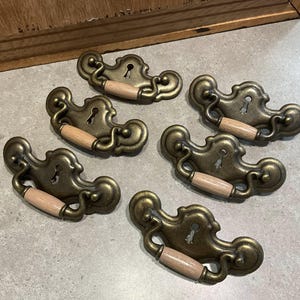 May include: Six antique brass drawer pulls with keyhole details and light wood handles. The ornate pulls have a vintage aesthetic, ideal for furniture restoration or adding a classic touch to cabinets and drawers.