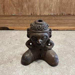 Pode incluir: Estatueta de cerâmica castanha escura que retrata uma figura sentada com um adorno de cabeça detalhado e traços faciais. A figura está ajoelhada com as mãos perto do peito. A escultura tem uma aparência desgastada, sugerindo uma origem antiga ou vintage.
