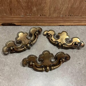 May include: Three antique brass drawer pulls with a cloud-like design. The pulls have a curved handle and decorative detailing. The brass has a warm, aged patina, suggesting vintage or antique hardware. These pulls are suitable for furniture restoration or adding a vintage touch.