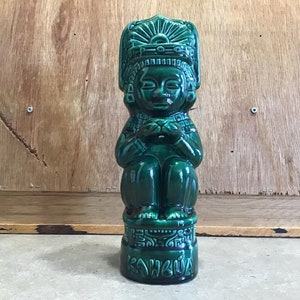 May include: Green ceramic figurine of a seated figure wearing a headdress with the word "Kahuna" on the base.