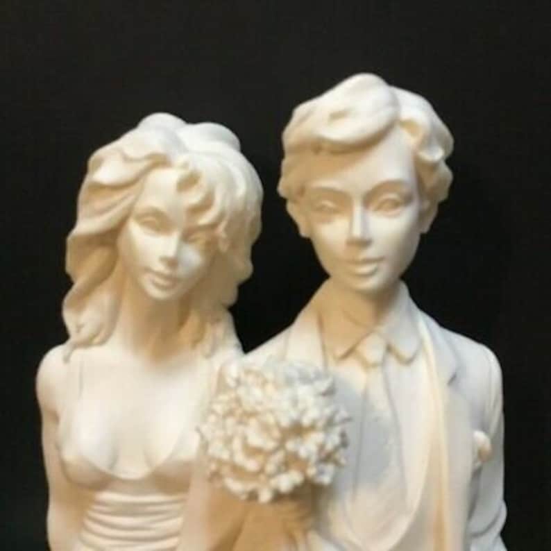 May include: White ceramic figurine of a bride and groom. The bride is wearing a strapless gown and the groom is wearing a tuxedo. They are holding a bouquet of flowers.