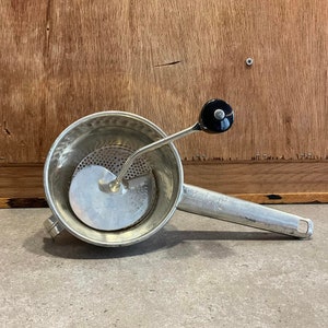 May include: A silver food mill with a black handle. The mill has a round bowl with a perforated disc and a long handle with a crank.