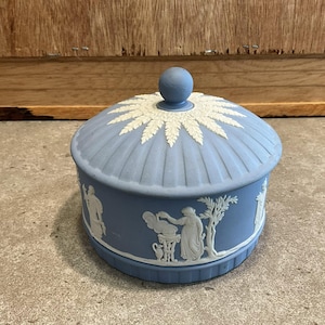 Puede incluir: Una caja de baratijas de cerámica azul y blanca con tapa abovedada. La tapa presenta un diseño de hojas blancas. La caja tiene figuras en relieve blancas alrededor de los lados.