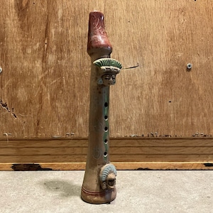 Pode incluir: Flauta de cerâmica marrom com bocal marrom avermelhado e entalhes decorativos. A flauta apresenta rostos esculpidos com cocares e detalhes verdes. O instrumento possui vários orifícios para os dedos e uma base alargada.