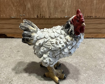 Peut inclure: Figurine décorative représentant un coq. Le coq a des plumes blanches et noires, une crête et des barbillons rouges. La figurine est posée sur une surface grise.