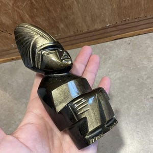 Pode incluir: Uma figura de obsidiana preta e dourada de uma pessoa com um chapéu. A figura é esculpida em estilo geométrico e tem um acabamento brilhante.