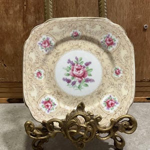 Op de afbeelding: Een vierkant bord in vintage-stijl met een bloemenontwerp. Het bord heeft een crèmekleurige achtergrond met een herhalend patroon en roze rozen en paarse bloemaccenten. Het bord wordt gepresenteerd op een decoratieve goudkleurige standaard.