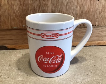 Op de afbeelding: Witte keramische mok met een rood Coca-Cola-logo en de tekst "Drink Coca-Cola in Bottles".