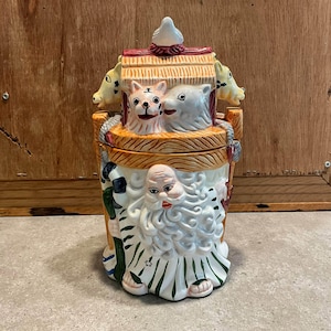 May include: A ceramic cookie jar featuring a sculpted figure with a long white beard and green robes. The lid is shaped like a wooden structure topped with a bird, flanked by two animal heads, and includes a tiger and a white animal face.