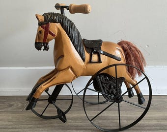 May include: A vintage wooden horse tricycle with a tan body, black mane, and red bridle accents. The horse has a black saddle and a brown tail. The tricycle features black metal wheels and a wooden handlebar.