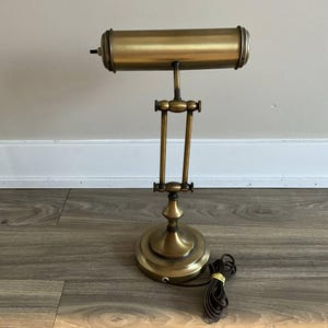 May include: A brass-toned desk lamp with an adjustable arm and a cylindrical shade. The lamp has a round base and a cord with a yellow band. The lamp is designed in a classic, vintage style.