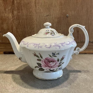 May include: White porcelain teapot with a large pink rose and floral design. The teapot has a curved spout, ornate handle, and a lid with a small knob. A gold trim accents the base and handle.