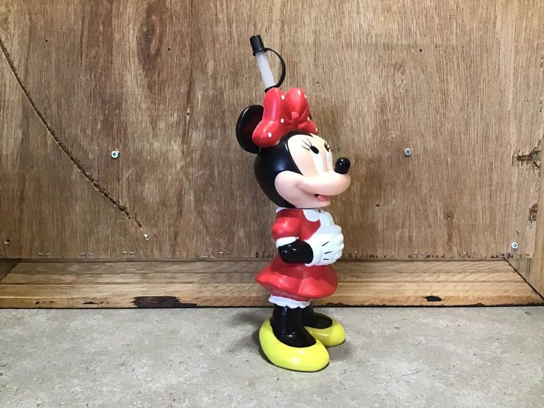 May include: A Minnie Mouse-shaped tumbler with a black head, red bow, and red dress. The tumbler has a black straw and yellow shoes. The design is a classic Disney character.