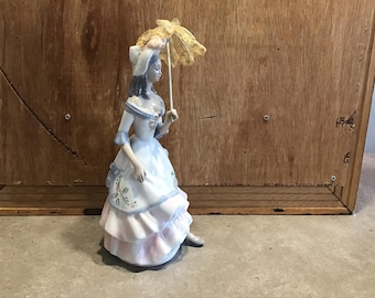 May include: A porcelain figurine of a woman in a white and pink dress holding a yellow and white umbrella.