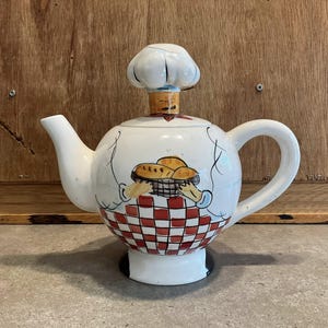 Peut inclure: Théière en céramique blanche avec un couvercle en forme de toque de chef. La théière a un corps rond avec un motif à carreaux rouges et blancs à la base et une image peinte de pain dans un panier. La poignée et le bec sont blancs.