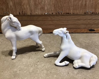 May include: Two white porcelain horse figurines. One horse is standing with its head turned back, while the other is lying down with its head turned to the side.