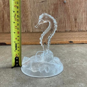 May include: A clear glass seahorse figurine with a textured surface. The seahorse is standing on a base with a textured surface that resembles a bed of seashells.