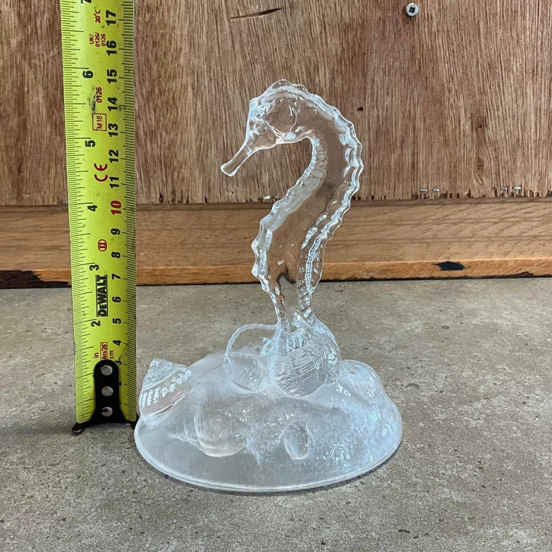 May include: A clear glass seahorse figurine with a textured surface. The seahorse is standing on a base with a textured surface that resembles a bed of seashells.