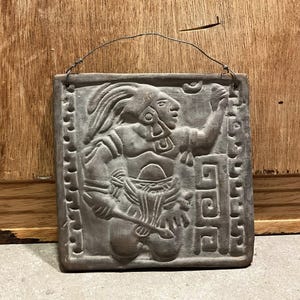 Op de afbeelding: Een grijze, rechthoekige reliëfsculptuur die een figuur in profiel afbeeldt, mogelijk Maya of Azteeks. De figuur is versierd met een hoofdtooi en sieraden, en houdt een voorwerp vast. De sculptuur heeft een draad om op te hangen en is omlijst door een gestippelde rand.
