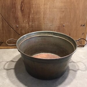 Puede incluir: Una olla grande, redonda, de cobre vintage con dos asas. La olla tiene una pátina verde y está sobre una superficie de madera.