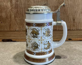 以下が含まれることがあります： 銀色の蓋とハンドルが付いた白いセラミックビールジョッキ。ジョッキは、金色の干支のシンボルと日付で装飾されています。金と茶色の装飾的なバンドがベースを囲んでいます。