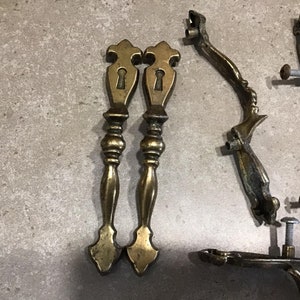 Puede incluir: Dos tiradores de puerta de armario de latón antiguo con detalles de ojo de cerradura. Los tiradores tienen un diseño decorativo y ornamentado.