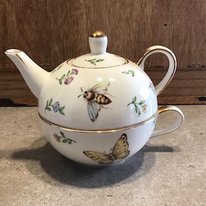 Puede incluir: Juego de tetera y taza de té de porcelana blanca con un diseño floral y de mariposa. La tetera tiene un borde dorado y un asa dorada. La taza tiene un borde dorado y un asa dorada.