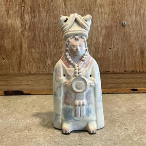 Pode incluir: Uma figurinha de cerâmica branca de uma pessoa usando um chapéu e uma túnica longa. A figurinha tem um desenho pintado em tons de azul, vermelho e marrom. A pessoa está segurando um objeto redondo em suas mãos.