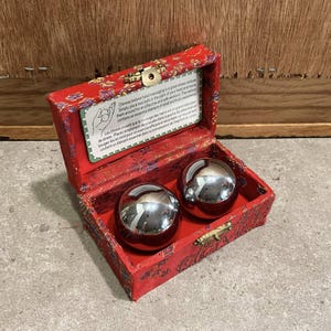 Peut inclure: Une boîte rouge à motifs avec un fermoir doré contient deux boules de massage argentées et brillantes. La boîte contient une petite carte avec du texte sur le massage chinois des mains. Les boules sont pour le soulagement du stress.