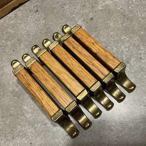 May include: Six wooden cabinet pulls with brass accents. The pulls have a rectangular wooden body with brass caps and feet. The wood grain is visible, and the brass has a slight sheen. The pulls are arranged diagonally on a gray surface.